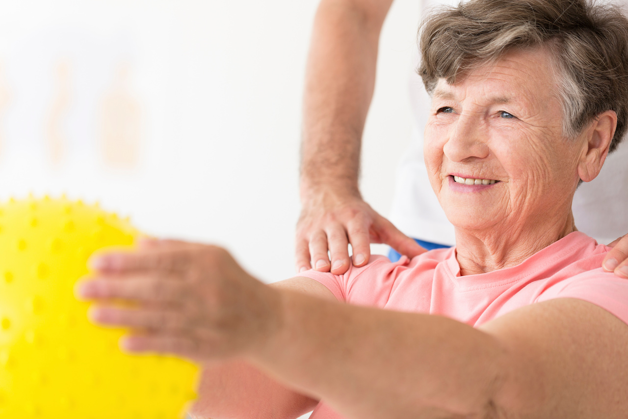 A close-up portrait of an elderly woman holding a ball in physiotherapy with doctor's hands on her shoulders